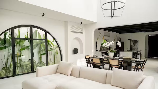 Una sala de estar moderna con un sofá blanco, una mesa de comedor y sillas, un ventanal con plantas, y una cocina al fondo.