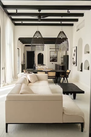A modern living room with a sofa, dining table, and kitchen area in the background. - Luxurious 3 BR villa, 100 meters from the Beach (Seseh)