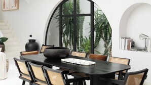 Uma sala de jantar com uma mesa redonda, cadeiras pretas e um espelho grande refletindo plantas.