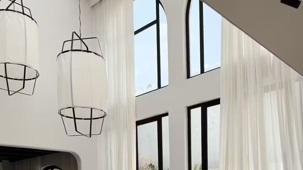 Ampla sala de estar com um sofá seccional grande, uma mesa de centro e uma poltrona de couro. Há grandes janelas com cortinas leves, uma área de jantar com mesa e cadeiras, e uma estante.