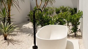 Ein modernes Badezimmer im Freien mit einer großen weißen Wanne, umgeben von kleinen Pflanzen und wolkenlosem Himmel.