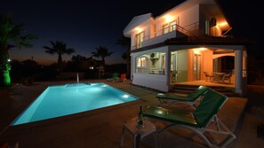 Una casa de dos pisos con piscina, mobiliario exterior y una entrada bien iluminada.