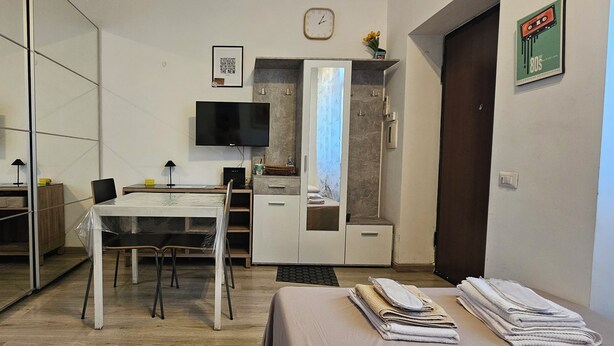 A modern hotel room with a glass partition, a dining table, a TV, a wardrobe, and a bed with neatly folded towels.