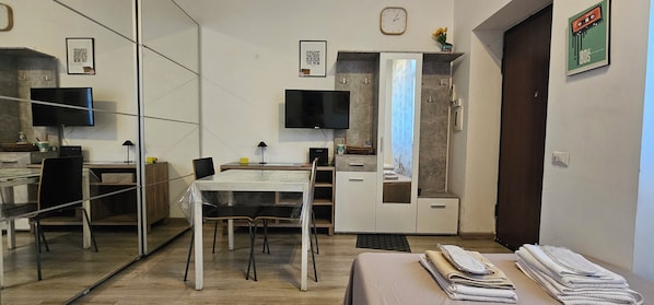 A modern hotel room with a glass partition, a dining table, a TV, a wardrobe, and a bed with neatly folded towels.