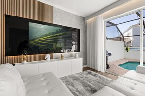 Ein modernes Wohnzimmer mit großem Flachbildfernseher, weißen Sofas, einer Wandpaneele aus Holz und Blick auf einen Swimmingpool.