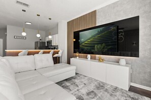 Ein modernes Wohnzimmer mit einem Ecksofa, einem flachbildfernseher an der Wand, einer Küche im Hintergrund und einem großen Fenster.