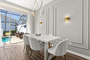 Ein moderner Essbereich mit einem weißen Tisch und Stühlen, einer Glastür, die zu einem Außenpool führt, und dekorativen Wandleuchten.