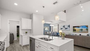 Cocina moderna con armarios blancos, una isla central con lavamanos, iluminación colgante y un comedor con vista al exterior.