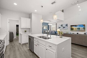 A modern kitchen with white cabinetry, a central island with a sink, pendant lighting, and a dining area with a view of the outdoors. - Last Minute Disney Getaway! Luxury Villa with Private Pool. Book Now! #155727 (Orlando)