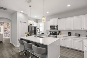 A modern kitchen with white cabinetry, a central island with bar stools, a stainless steel refrigerator, a microwave, and an oven. - Last Minute Disney Getaway! Luxury Villa with Private Pool. Book Now! #155727 (Orlando)