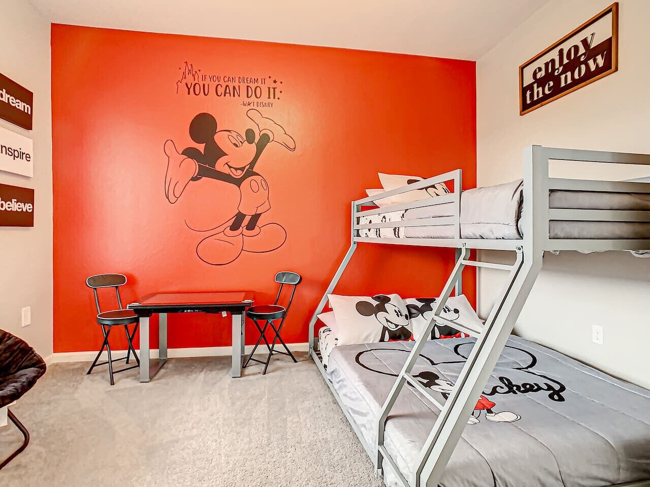 Un lit superposé avec une parure de lit aux couleurs de Mickey Mouse, un bureau avec des chaises et une citation inspirante sur le mur.