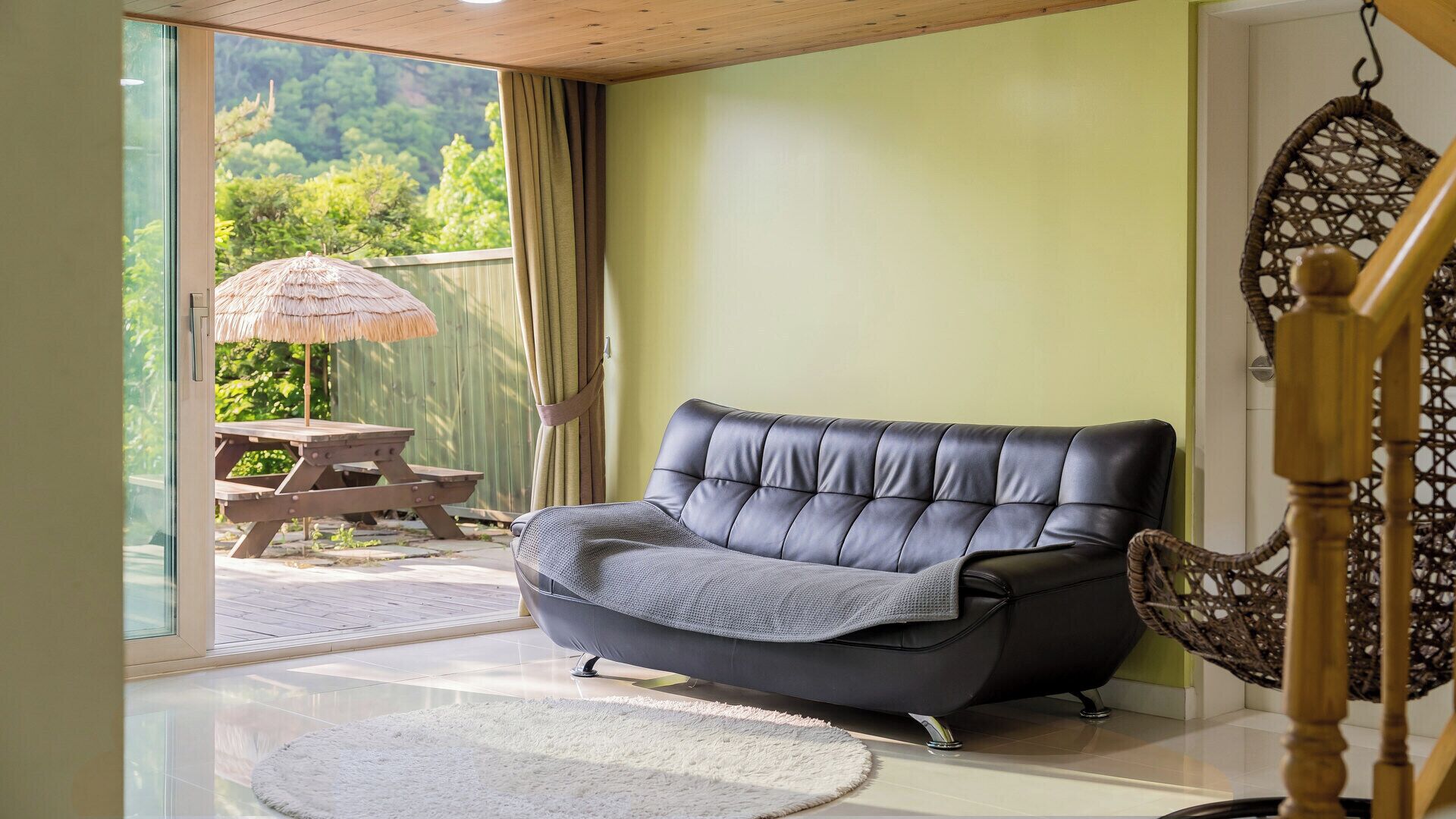 A modern living room with a leather sofa, a wooden table with an umbrella, and a view of greenery through a glass door.