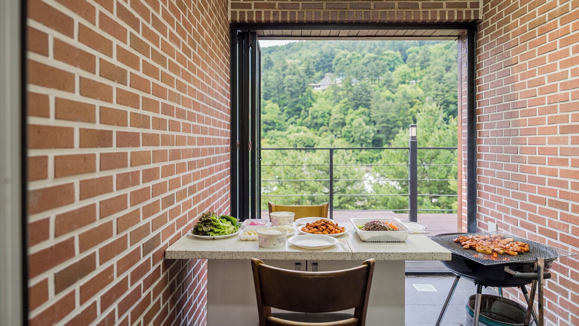 A balcony with a brick wall, a table with food, and a grill.