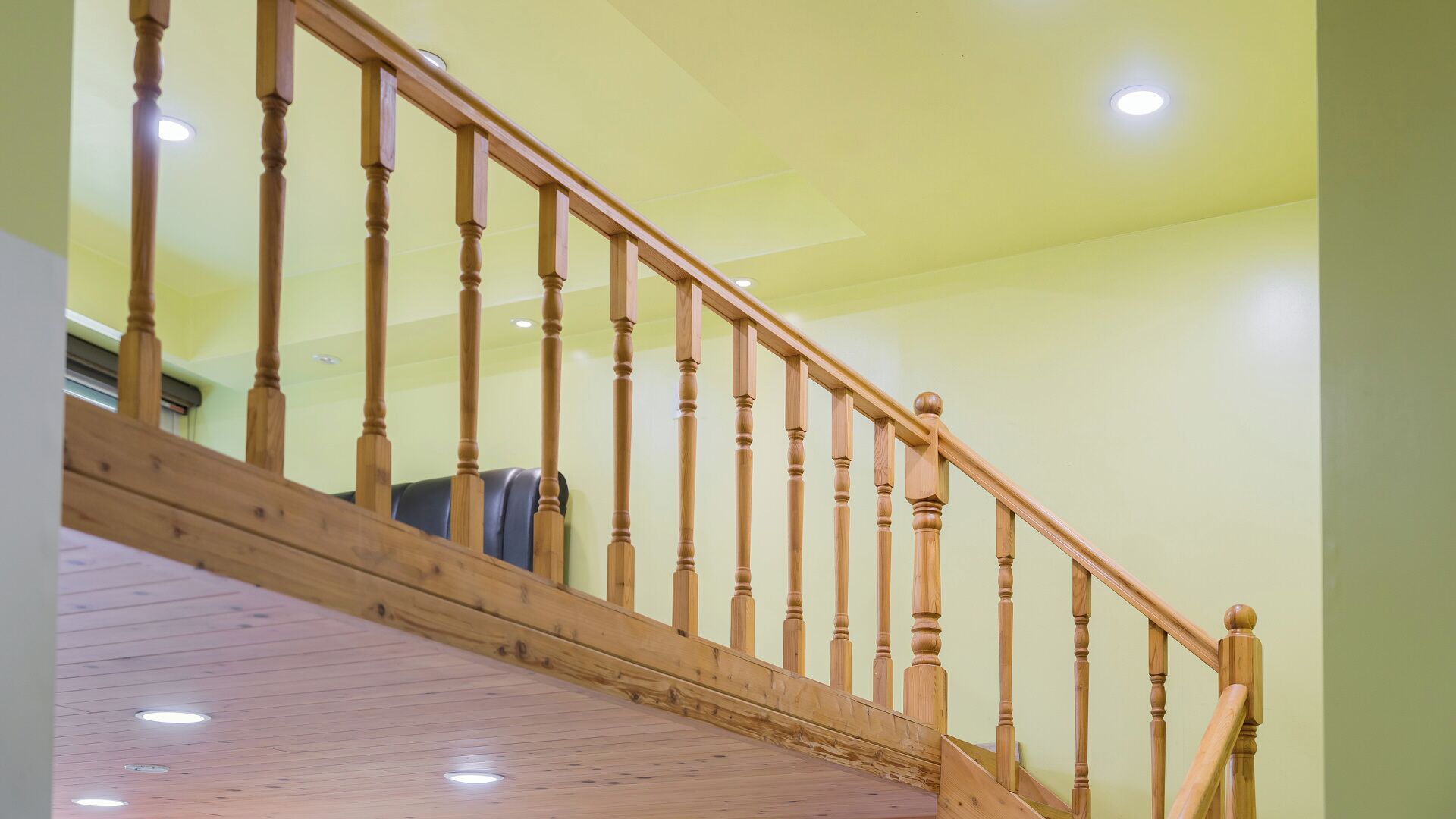 Una escalera de madera con barandilla, una lámpara en el techo y vista de una habitación con una silla.
