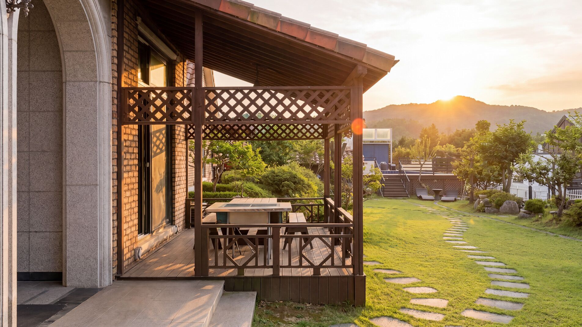 A covered outdoor seating area with a wooden pergola, a paved walkway, and a view of a building and hills in the background.