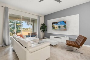 Ein modernes Wohnzimmer mit einem weißen Sofa, einem braunen Sessel, einer an der Wand befestigten TV-Anlage über einer weißen Konsole und einem Balkon mit einer Sitzbank.