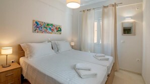 A neatly made bed with white linens, a bedside lamp, a colorful abstract painting on the wall, and a window with curtains.
