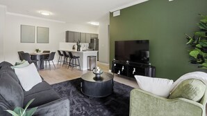 A modern living room with a black coffee table, a green armchair, a dining area with a kitchen in the background, and a flat-screen TV mounted on the wall. - Spacious 3-bedroom apartment - Close to Food and Fun! (Houston)