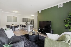 Ein modernes Wohnzimmer mit einem schwarzen Couchtisch, einem grünen Sessel, einem Essbereich und einer dahinterliegenden Küche. An der Wand ist ein Flachbildfernseher angebracht.