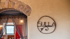 Un arc en briques mène à une pièce où se trouvent une table et des chaises, et un panneau circulaire portant le mot « SULLA » est fixé au mur.
