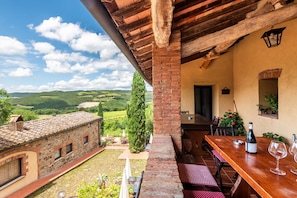 Une terrasse avec une table en bois, des chaises et une bouteille de vin. On peut admirer une vue sur des collines verdoyantes et un ciel dégagé.