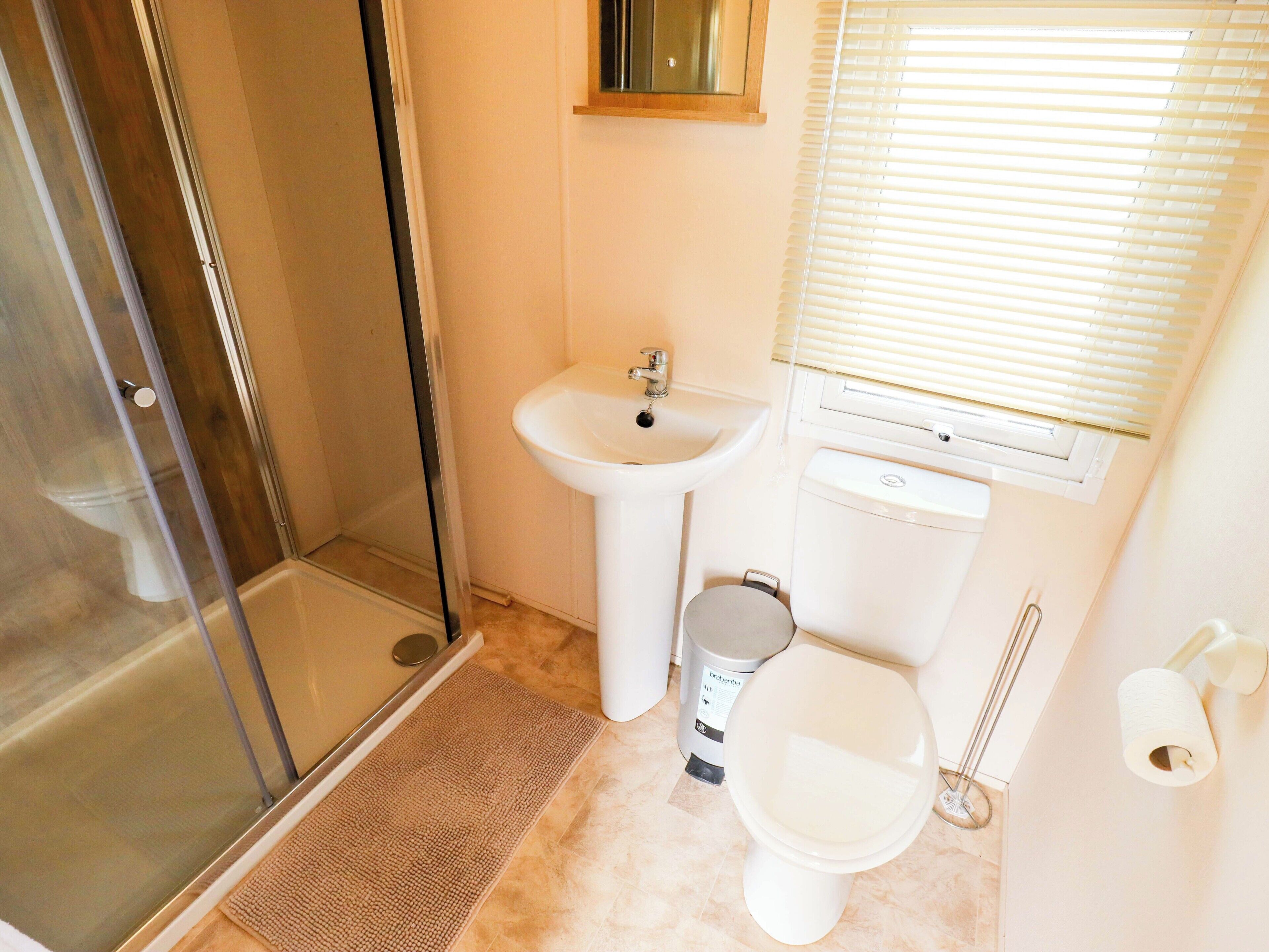 A bathroom with a glass shower enclosure, a pedestal sink, a toilet, and a window with blinds.