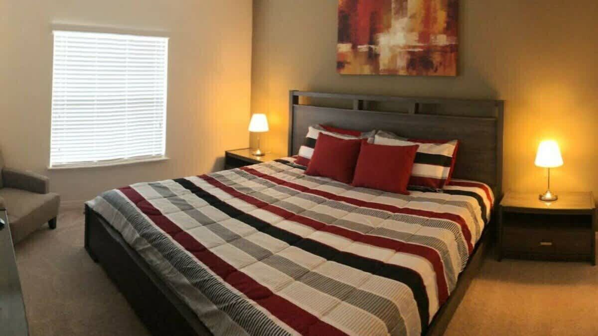 A bedroom with a large bed, two bedside tables with lamps, a window with blinds, and a painting on the wall.