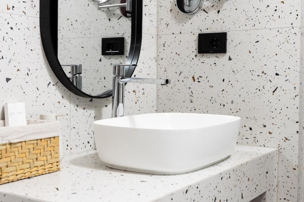 Un baño con salpicadero de azulejos blancos con motas, un lavamanos blanco y un espejo con marco negro.
