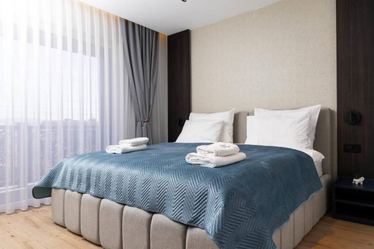 Moderni hotellihuone, jossa on suuri sänky, yöpöytä ja näkymä kaupunkiin ohuiden verhojen läpi.