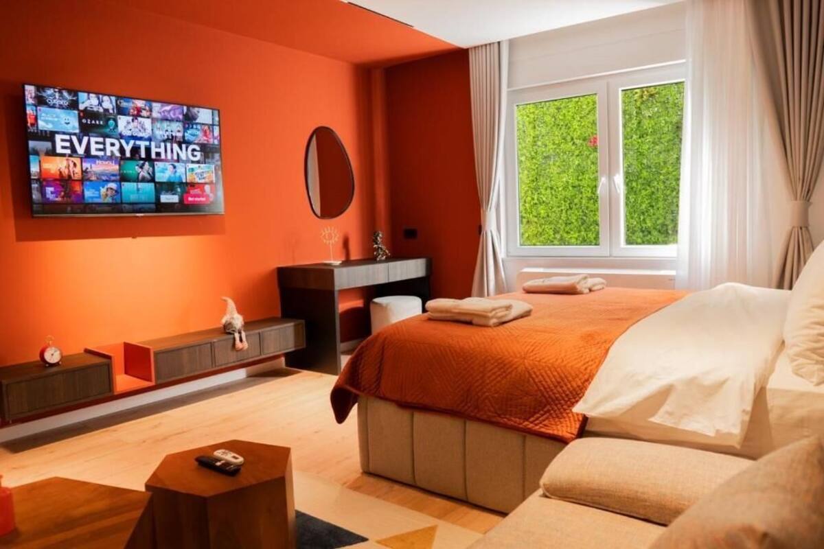 Moderni hotellihuone, jossa on suuri televisio, jolta näkyy useita kanavia, oranssi peitto sängyllä, yöpöytä kaukosäätimellä ja ikkuna, josta avautuu näkymä vehreään maisemaan.