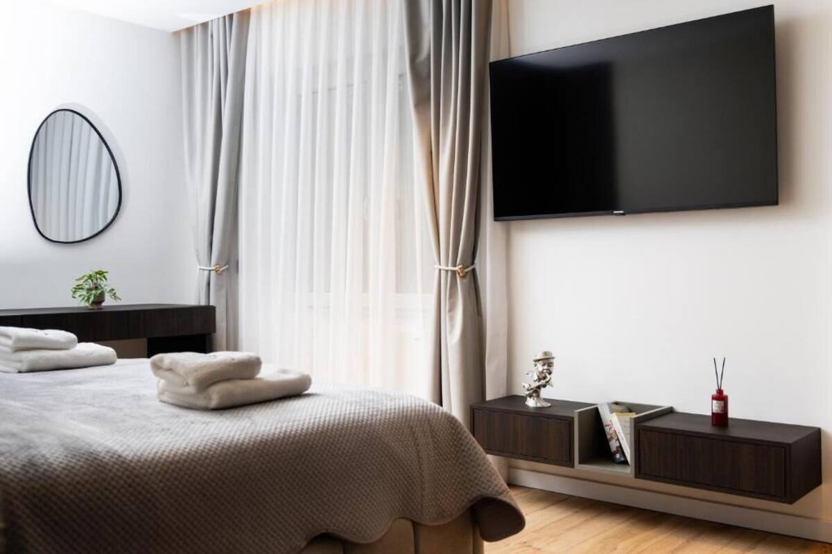 Moderni hotellihuone, jossa on suuri sänky, seinälle asennettu taulutelevisio, yöpöytä lampun kanssa ja ikkuna, jossa on verhot.