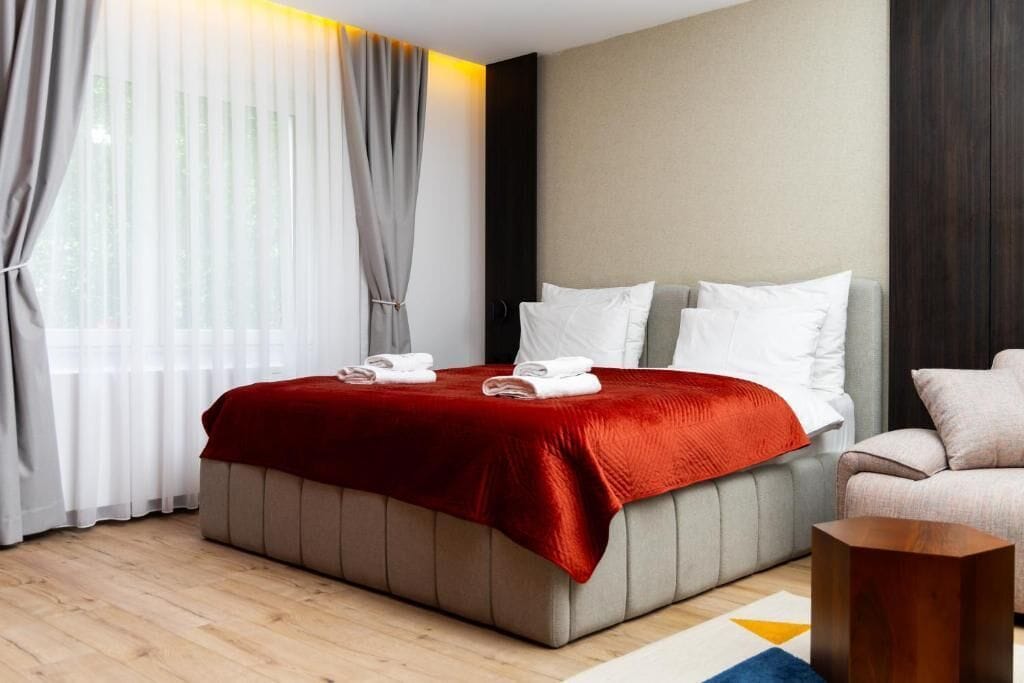 Une chambre d’hôtel moderne, équipée d’un lit, d’un canapé, d’une table d’appoint en bois et de rideaux légers.
