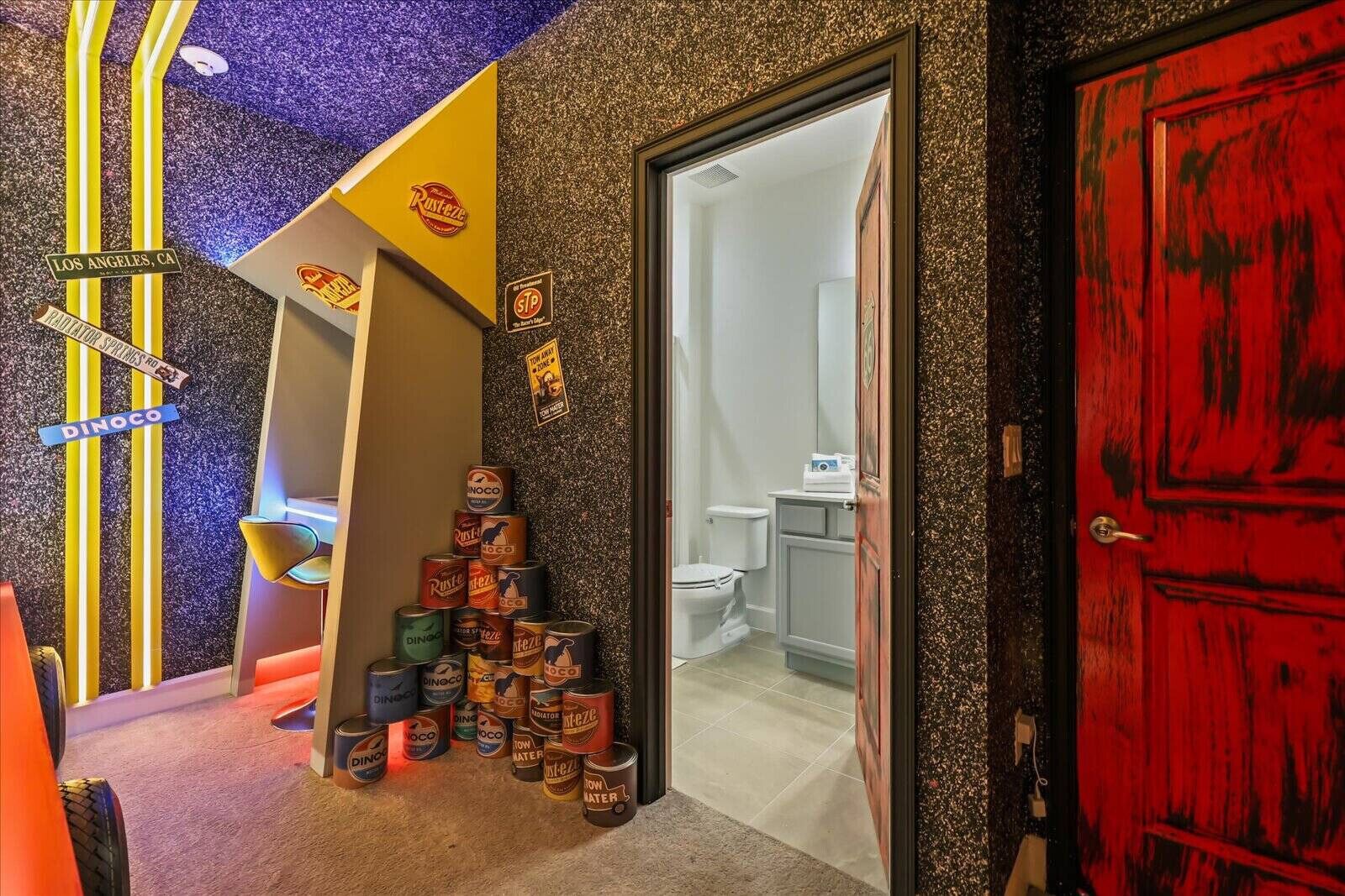 Habitación de hotel con una decoración original que incluye latas de bebidas, una puerta roja y una vista parcial de un baño.