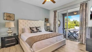 Une chambre à coucher comprenant un lit, deux tables de chevet, un ventilateur de plafond et une vue sur l’extérieur, accessible par une porte vitrée.