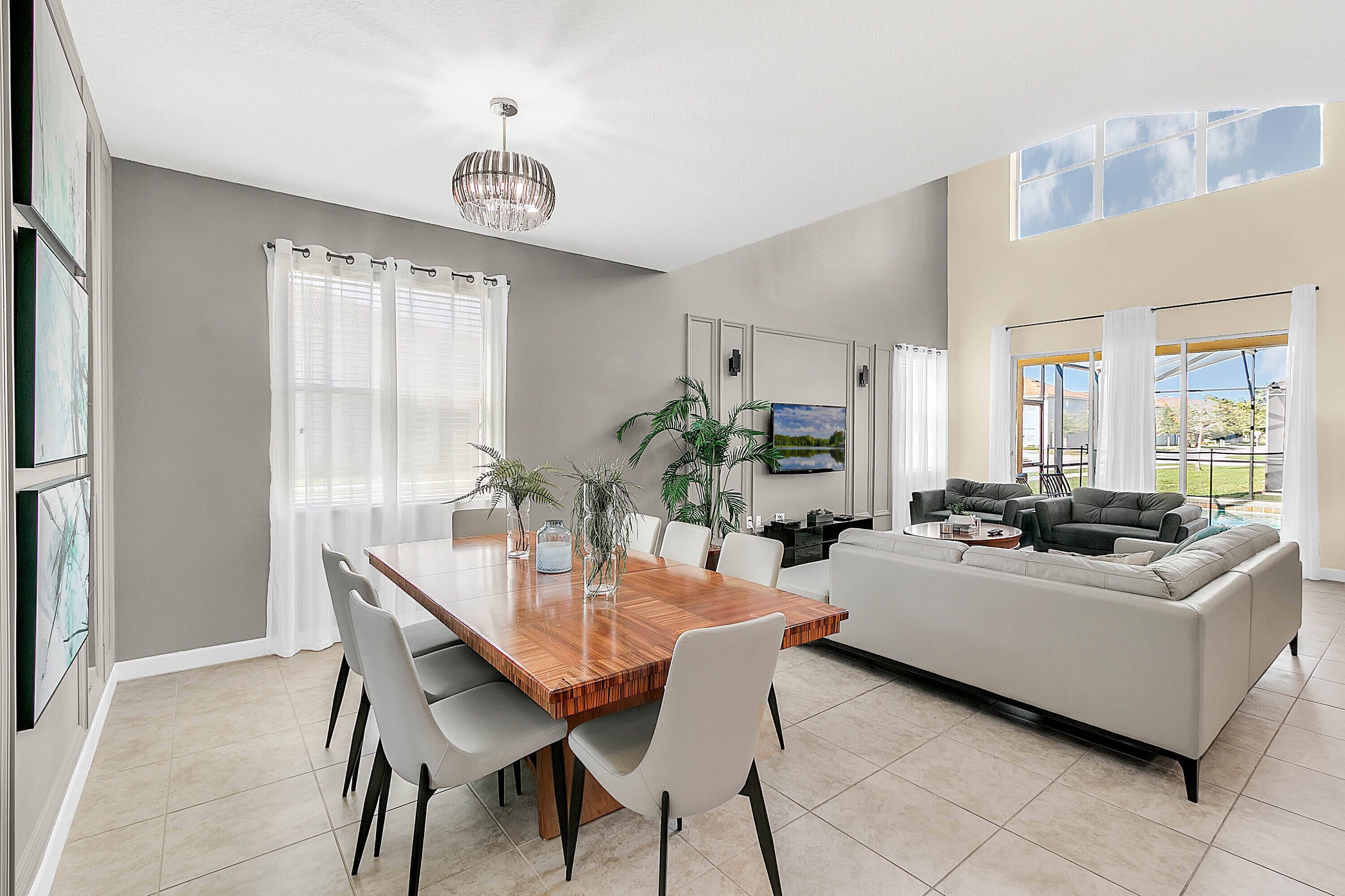 A spacious living area with a large wooden dining table, white chairs, a grey sofa, and a TV mounted on the wall.