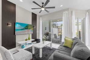 Ein modernes Wohnzimmer mit Deckenventilator, Flachbildfernseher, Glastisch, grauem Sofa und Blick auf eine Terrasse mit Grill.