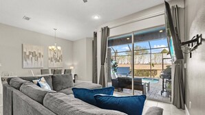 En moderne stue med en grå modulsofa, et spiseområde med bord og stole, samt en stor skydedør til en udendørs terrasse med pool og grill.