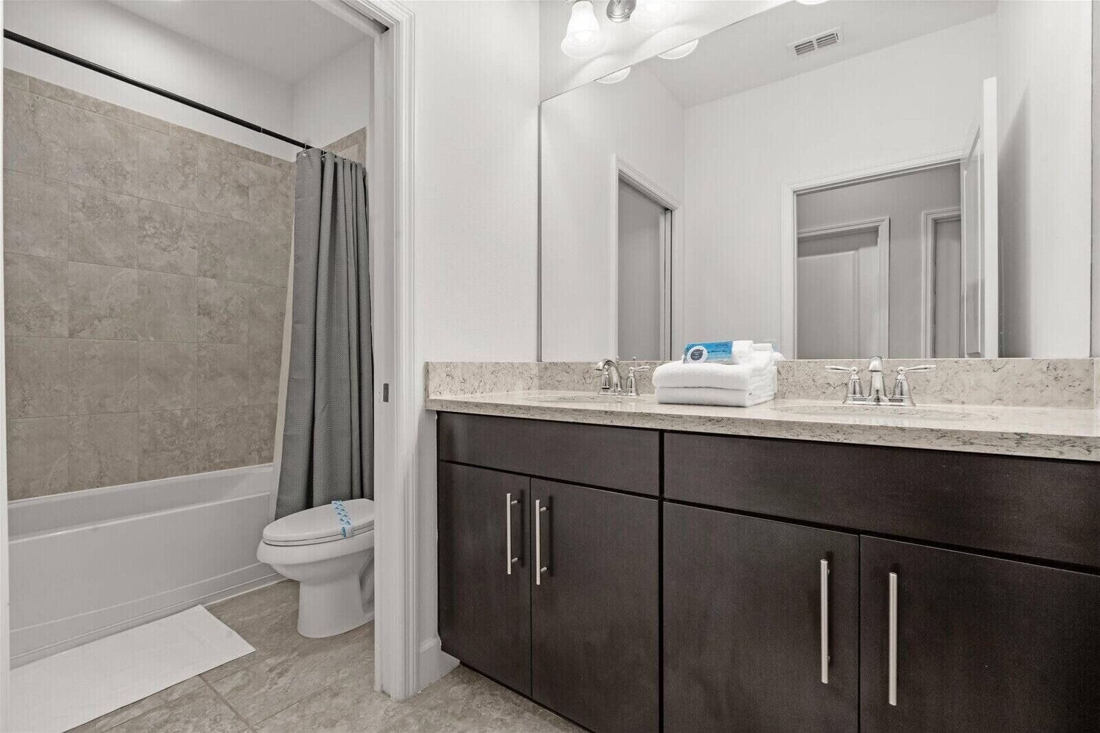 Une salle de bain avec une baignoire, des toilettes, des meubles sombres, un grand miroir et un plan de toilette avec des articles de toilette.
