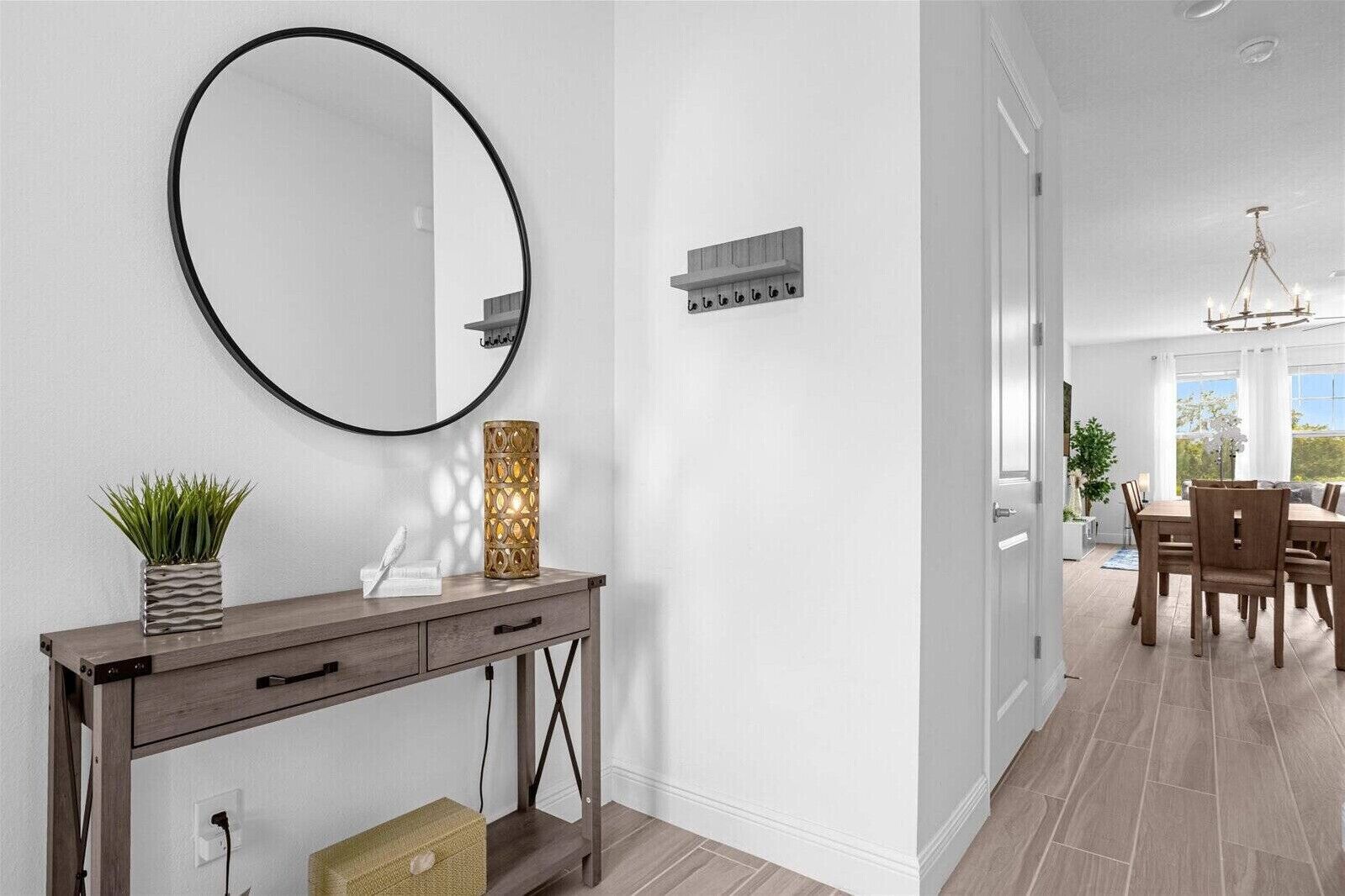 Un interior moderno con un espejo redondo, una consola con objetos decorativos, una lámpara de pared y una zona de comedor con luz natural.