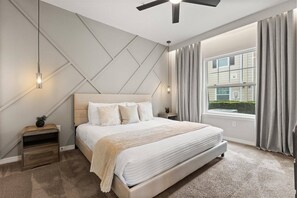 Ein Schlafzimmer mit Bett, Nachttisch, Deckenventilator, Fenster mit Vorhängen und Blick auf ein Gebäude.