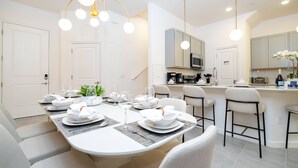 Un comedor moderno con una mesa larga preparada para una comida, una cocina al fondo y una zona de bar con taburetes.
