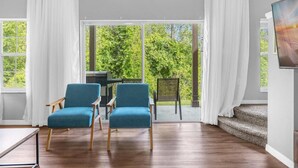 Una habitación con un gran ventanal corredizo que da a un balcón, dos sillas azules, una mesa de madera y un cuadro en la pared.