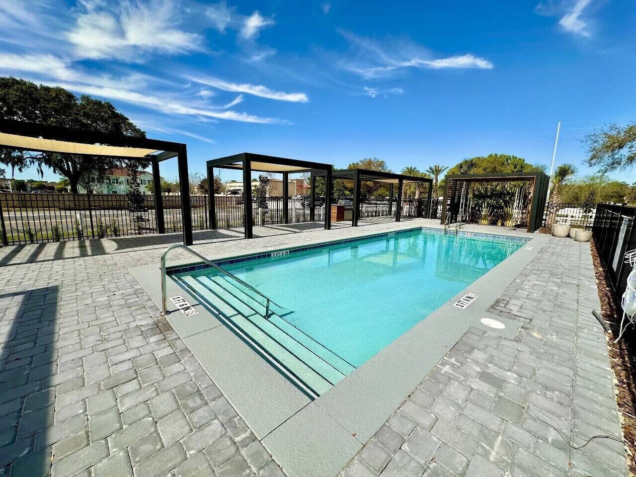 Une piscine rectangulaire, bordée d’une terrasse carrelée, entourée d’une clôture et agrémentée de zones ombragées.