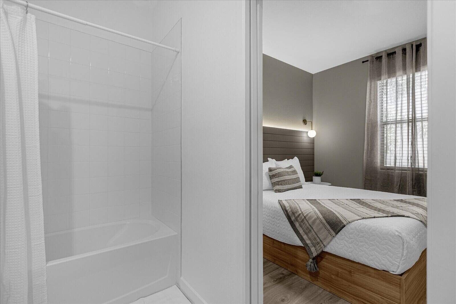 A modern bathroom with a shower, a bed with a headboard, a bedside table, and a window with curtains.