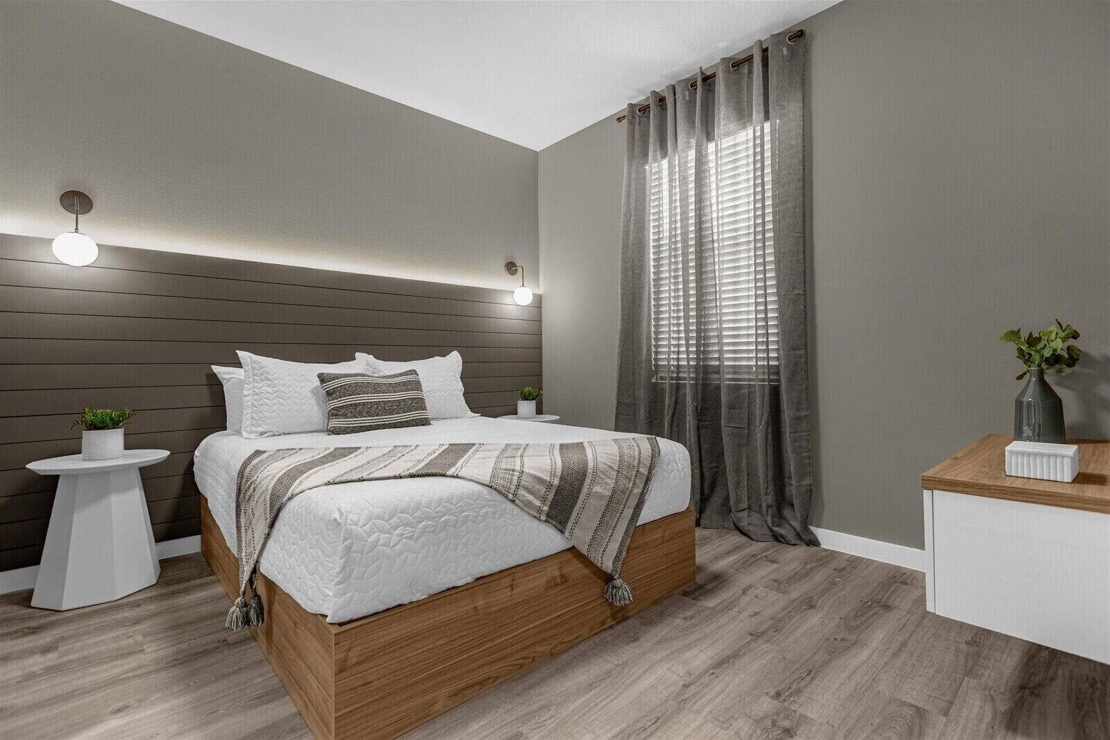 Une chambre moderne avec un lit en bois, une table de chevet agrémentée d’une plante, une commode et une grande fenêtre avec des rideaux.