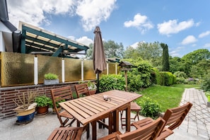 Een terras met een houten tafel en stoelen, een pergola en een tuin met diverse planten.