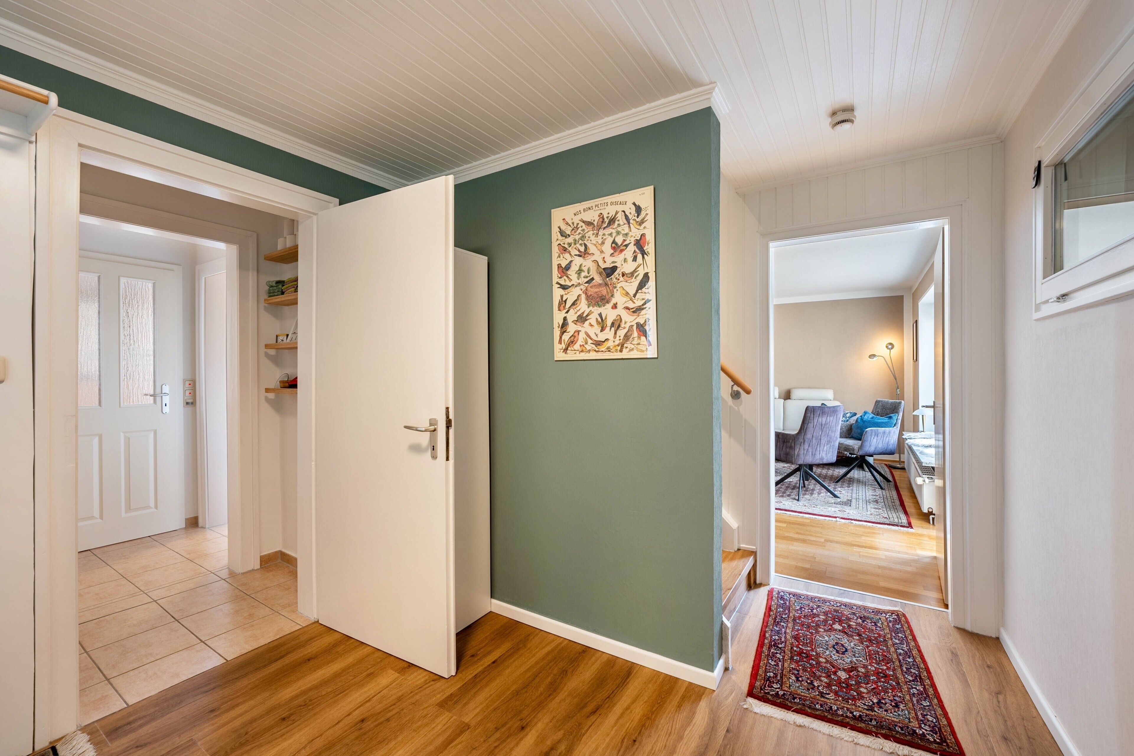 Un pasillo con suelo de madera, pared verde y una puerta que da a una habitación con una silla y una mesa.