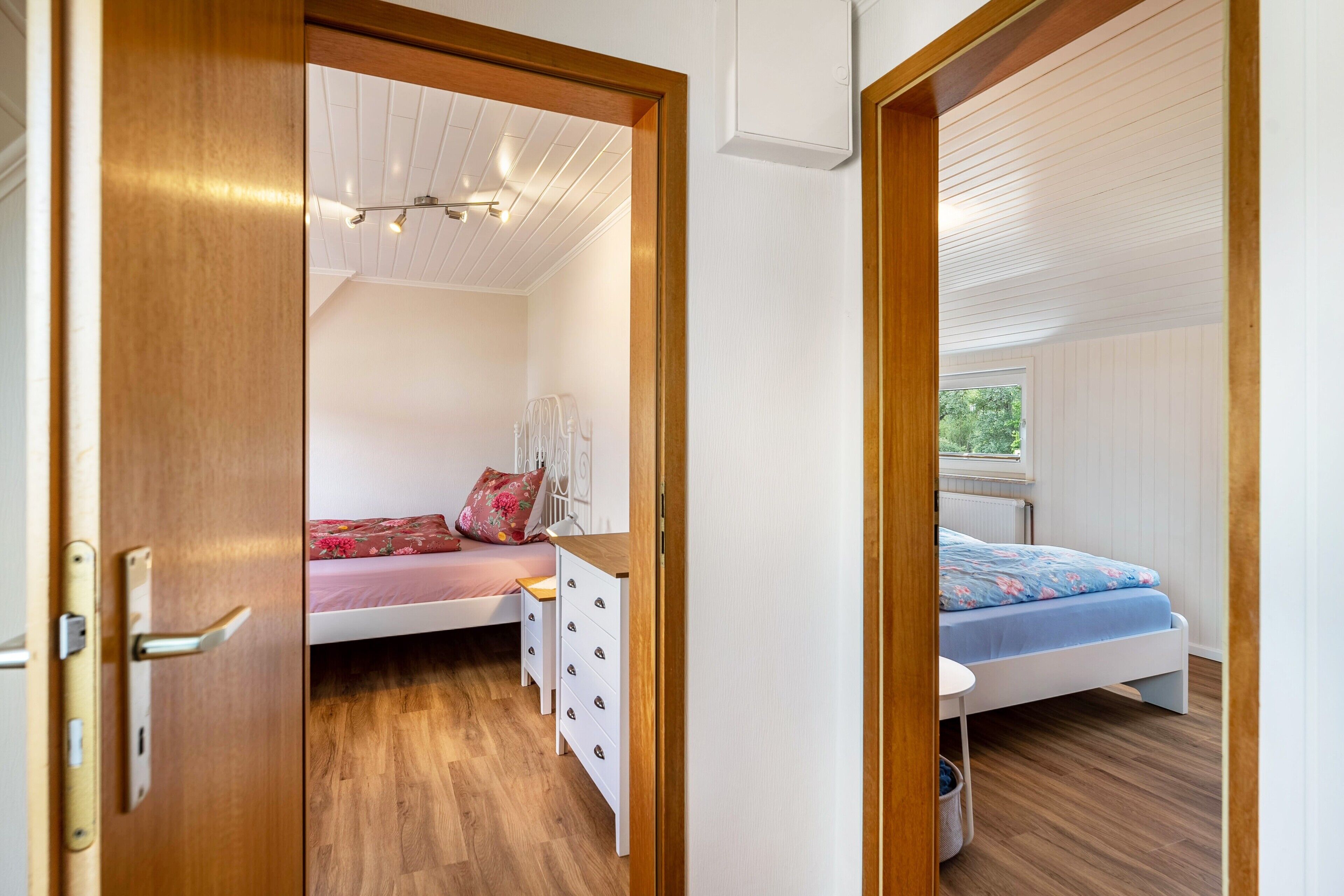 Un dormitorio con puerta de madera, cama con colcha estampada, cómoda blanca y ventana con vista a follaje.