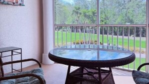 Un balcón con una mesa de vidrio y dos sillas de mimbre, con vista a un paisaje verde.