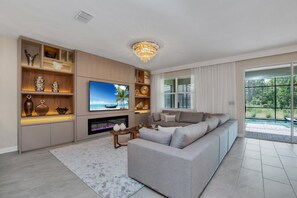 A modern living room with a large flat-screen TV, a fireplace, a sofa, and a view of a pool through large windows. - Last Minute Disney Getaway! Private Pool, Game Room & Themed Bedrooms. Book Now! #172197 (Orlando)